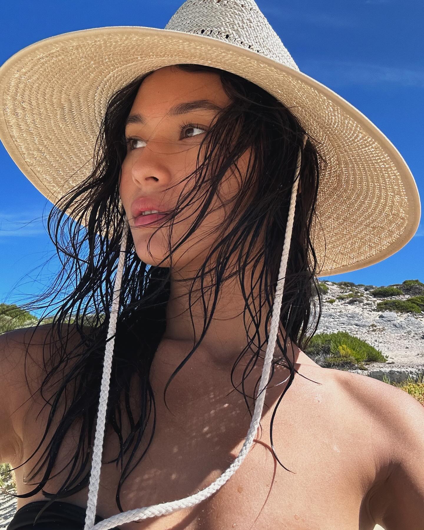 Close-up of a woman with dark hair wearing a straw hat and bikini top, posing on a sunny beach with a blue sky.