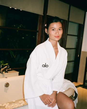 Asian woman in a white bathrobe sitting on a bathtub in an indoor bathroom with dark wood accents, and neutral tones at nighttime.