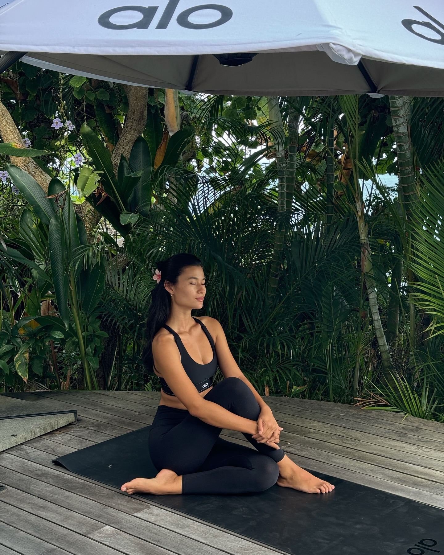 A woman in black athletic wear sits on a yoga mat outside, with lush tropical plants in the background and an umbrella overhead.