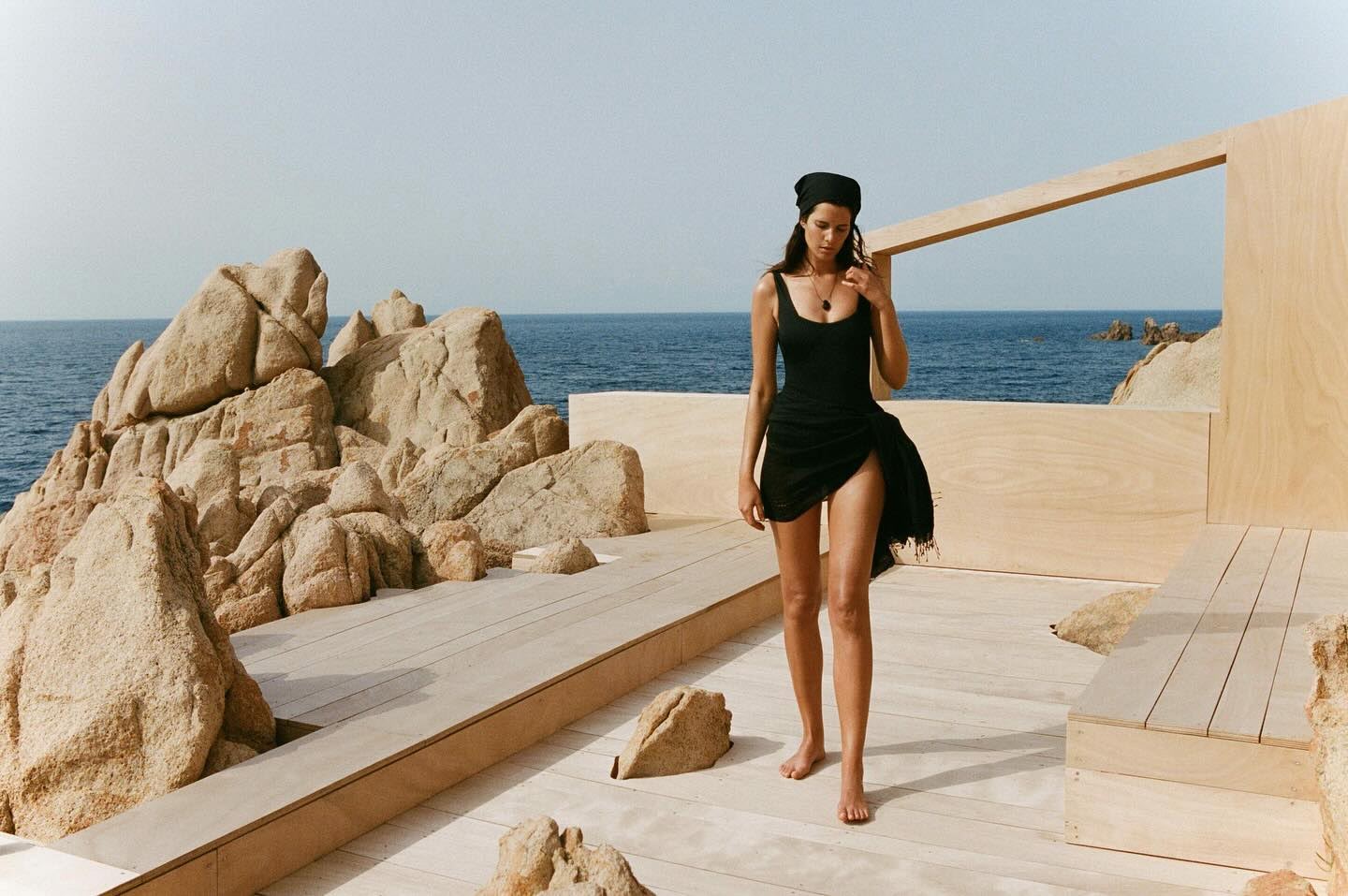 A woman in a black swimsuit and headscarf stands on a wooden platform near a rocky beach and ocean under a bright sky.