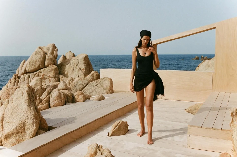 A woman in a black swimsuit and headscarf stands on a wooden platform near a rocky beach and ocean under a bright sky.