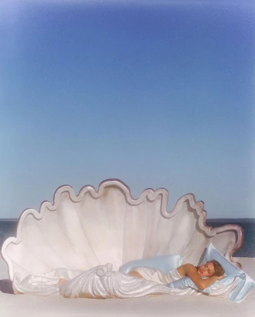 Woman resting inside a large sculpted seashell bed on a sandy beach against a clear blue sky