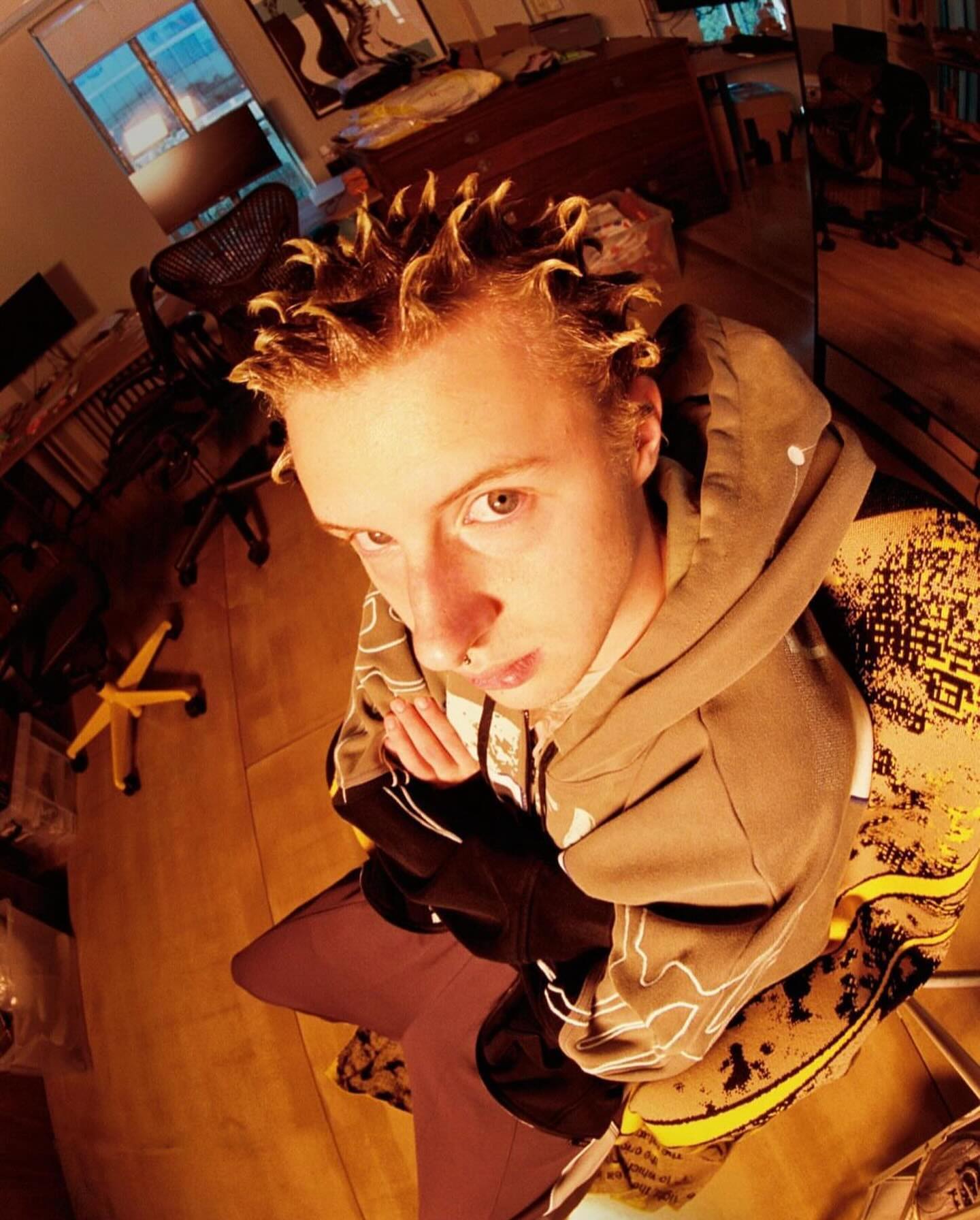 Indoor portrait of a young adult male in a hoodie, looking at the camera with a neutral expression, using a wide-angle, upward-tilted framing.