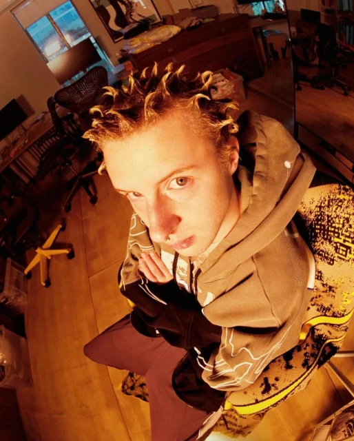 Indoor portrait of a young adult male in a hoodie, looking at the camera with a neutral expression, using a wide-angle, upward-tilted framing.