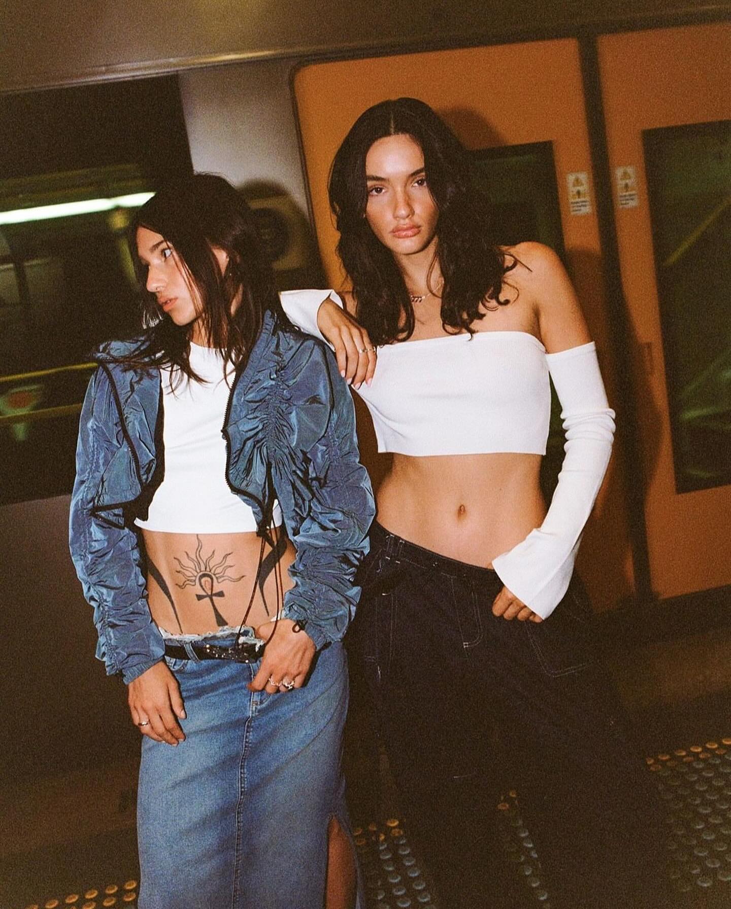 Two young women pose in modern fashion: crop tops, denim skirt, and jacket in an underground setting with cool, edgy style.