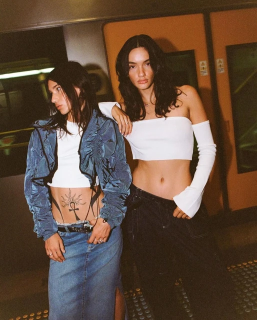 Two young women pose in modern fashion: crop tops, denim skirt, and jacket in an underground setting with cool, edgy style.