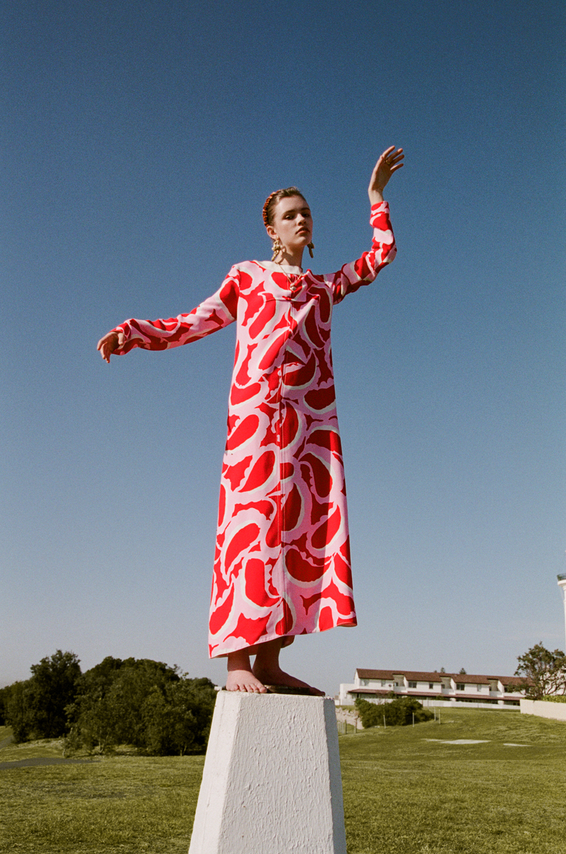 A woman in a pink and red dress stands on a plinth outdoors under a clear blue sky, posing with outstretched arms on a grassy lawn.