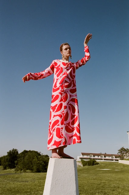 A woman in a pink and red dress stands on a plinth outdoors under a clear blue sky, posing with outstretched arms on a grassy lawn.
