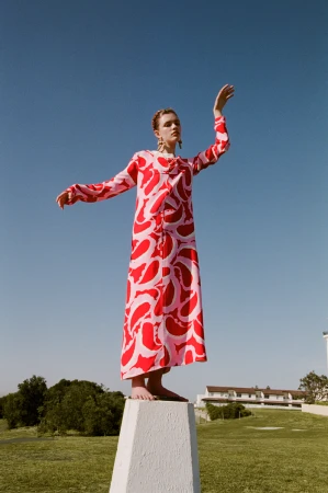 A woman in a pink and red dress stands on a plinth outdoors under a clear blue sky, posing with outstretched arms on a grassy lawn.