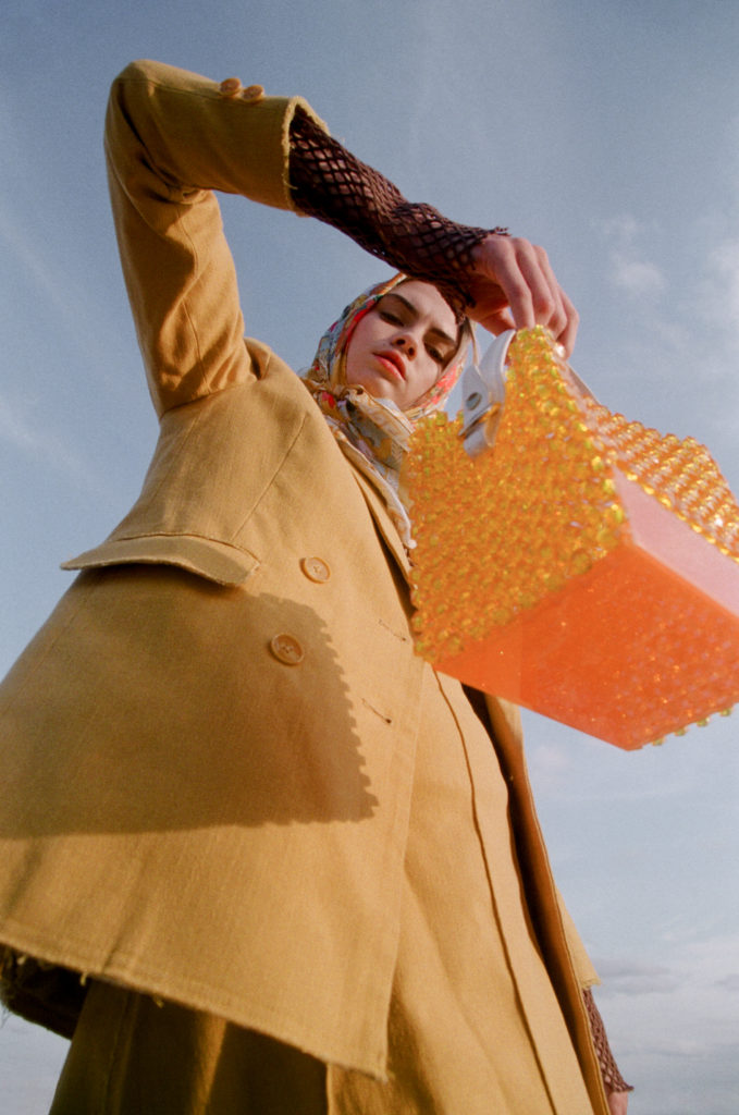 Fashionable woman posing in a mustard-colored suit and headscarf, holding a unique orange beaded bag under a bright, cloudless sky.