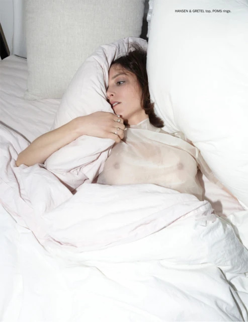 Woman lying in bed with white bedding and pillows, wearing a sheer top. Portrait-style shot with soft, diffused lighting. Fashion.