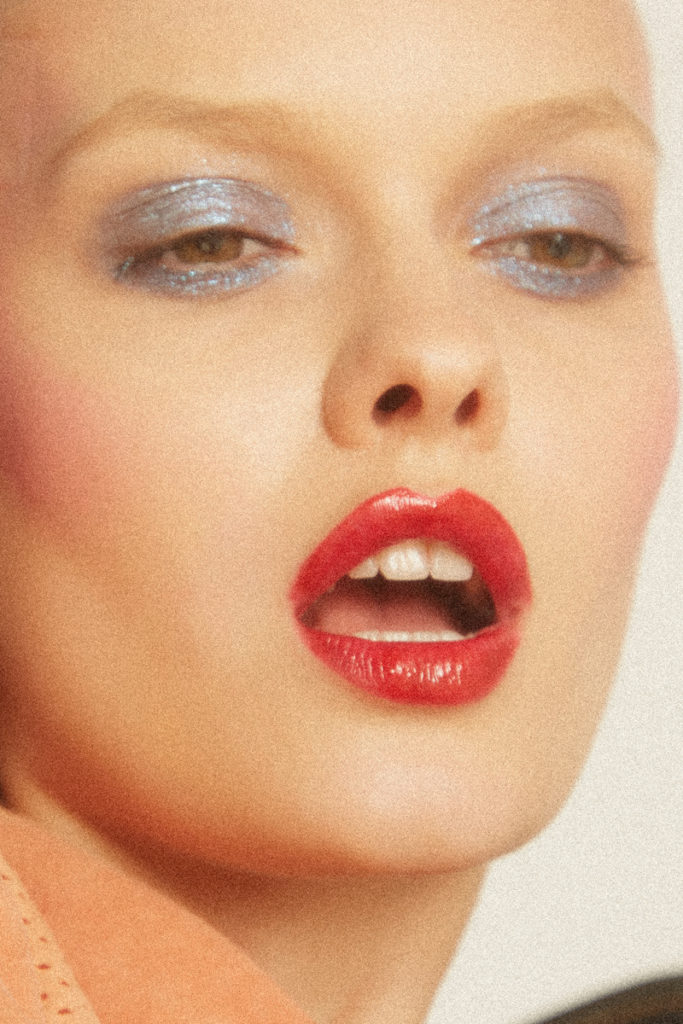 Extreme close-up shot of a fair-skinned woman with shimmery blue eye shadow and glossy red lips, gazing upward, against a neutral backdrop.