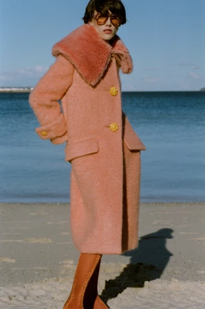 Adult female wearing a salmon-colored wool coat with matching boots on a beach with ocean and blue sky in the background on a sunny day.