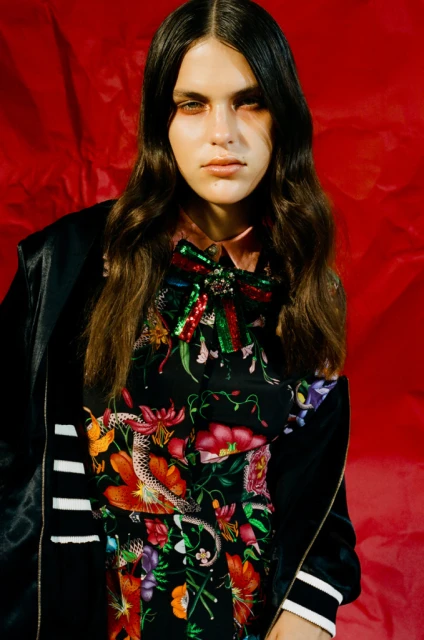 A woman with long brown hair poses in a black jacket and floral dress against a textured red background in a fashion editorial shot.