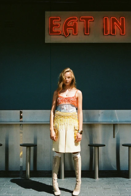 A woman in a colorful top and skirt stands beneath an 'EAT IN' neon sign with stools and a grey background, casting a shadow.