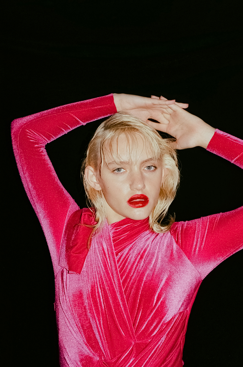 Close-up portrait of a fair-skinned young adult woman with blonde hair and red lipstick, wearing a pink velvet dress against black backdrop.
