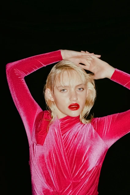 Close-up portrait of a fair-skinned young adult woman with blonde hair and red lipstick, wearing a pink velvet dress against black backdrop.