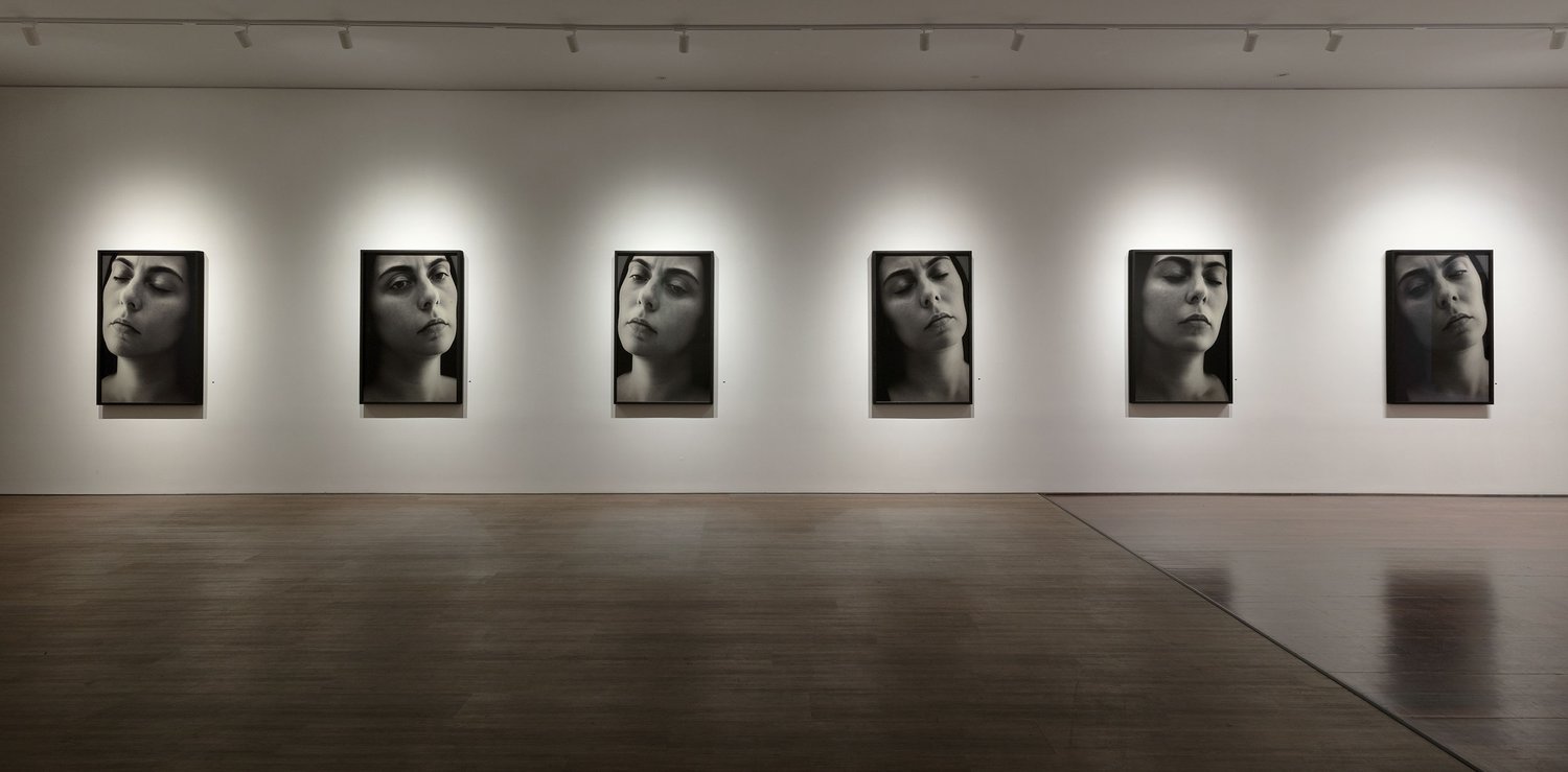 Six black-and-white portraits of the same woman displayed in a row in an art gallery with overhead lighting and wood flooring.