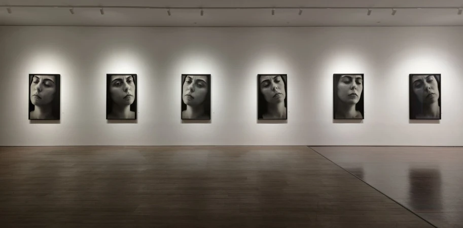 Six black-and-white portraits of the same woman displayed in a row in an art gallery with overhead lighting and wood flooring.