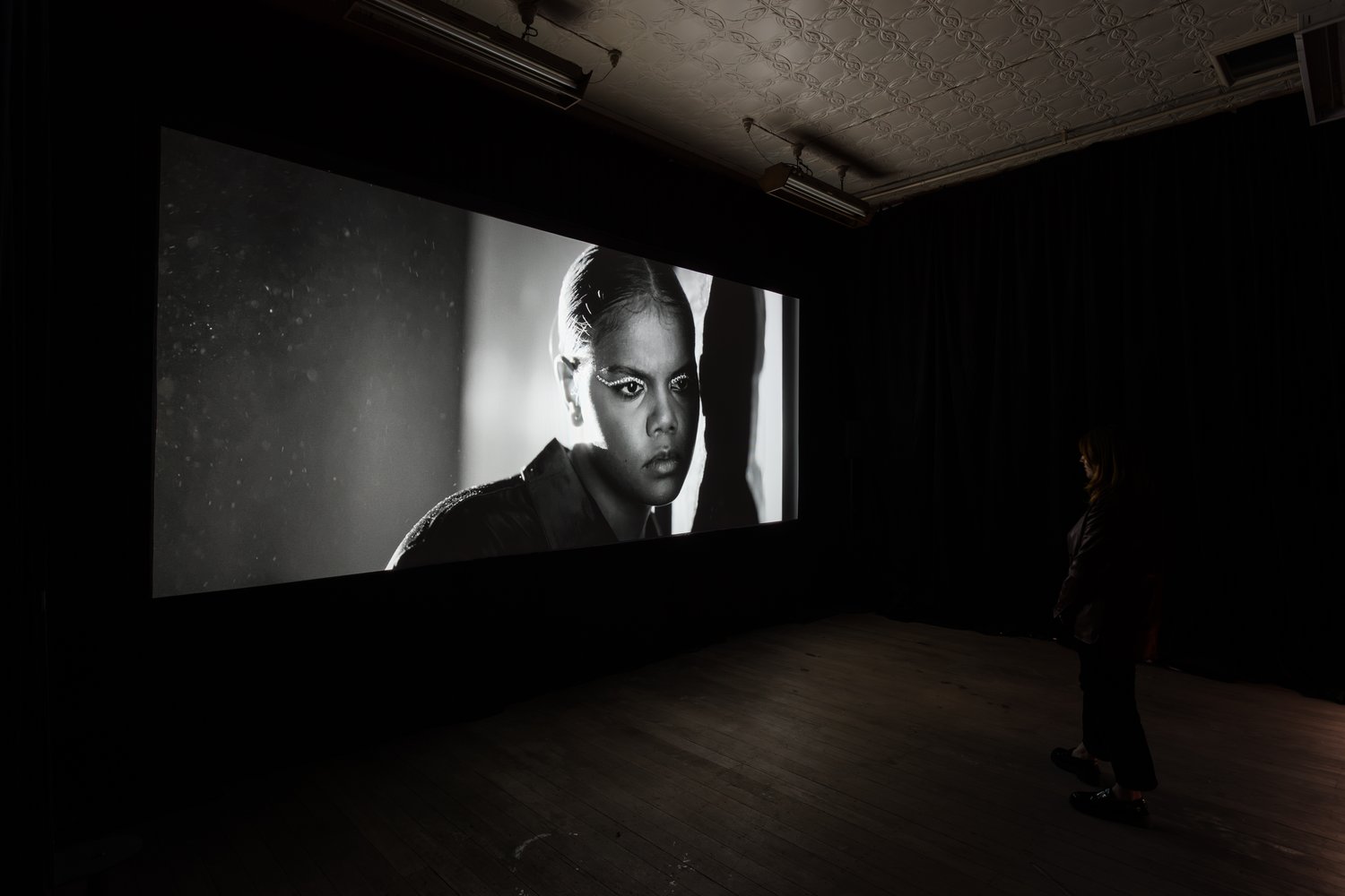 An adult female portrait with eye makeup, displayed on a large screen in a darkened room with a viewer, creating a monochromatic art exhibit.