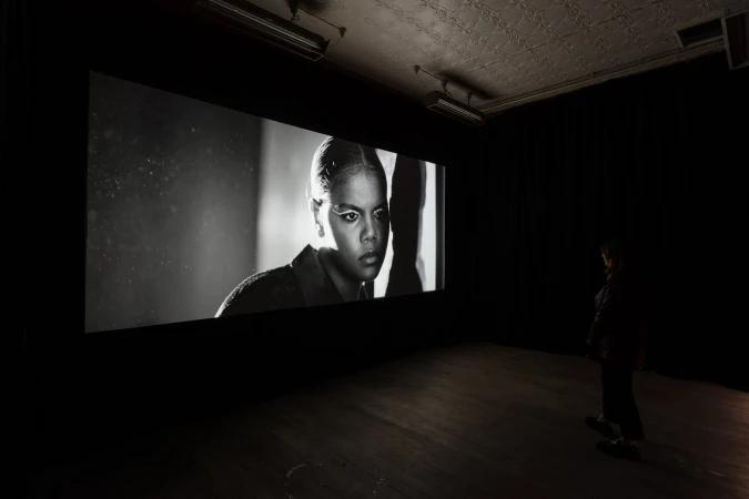 An adult female portrait with eye makeup, displayed on a large screen in a darkened room with a viewer, creating a monochromatic art exhibit.