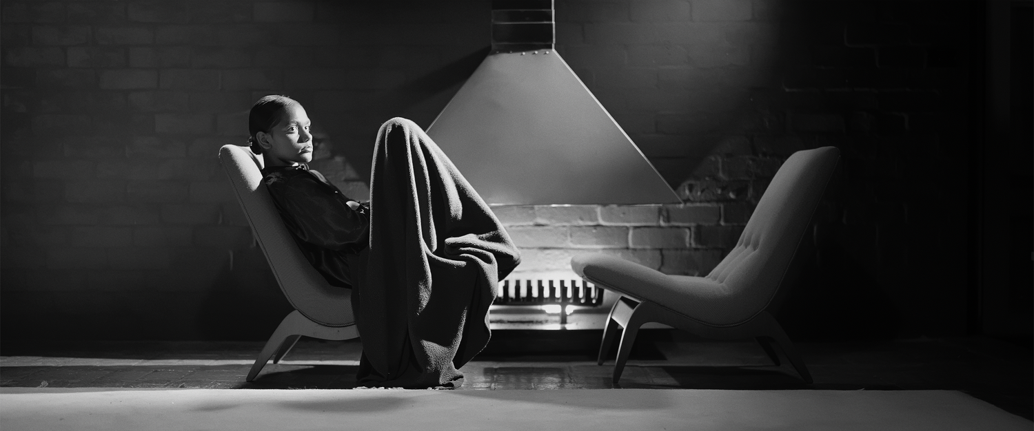 Black and white portrait of a person relaxing in a modern chair by a minimalist fireplace, showcasing interior design and comfort.