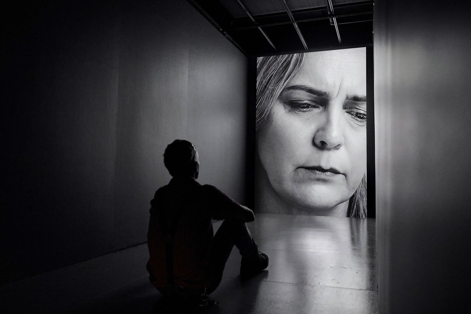 Monochromatic shot of a person sitting in a dark room looking at a portrait of a woman projected on a wall in front of them.