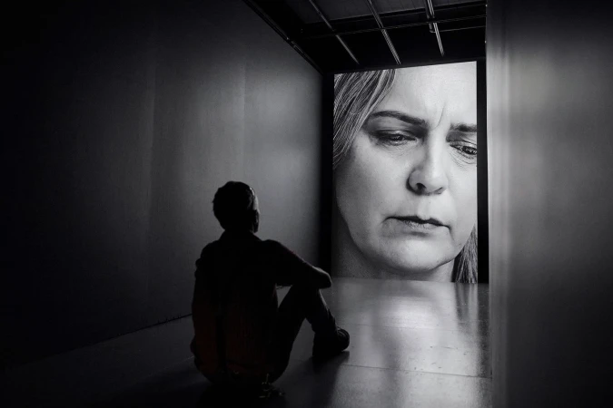 Monochromatic shot of a person sitting in a dark room looking at a portrait of a woman projected on a wall in front of them.