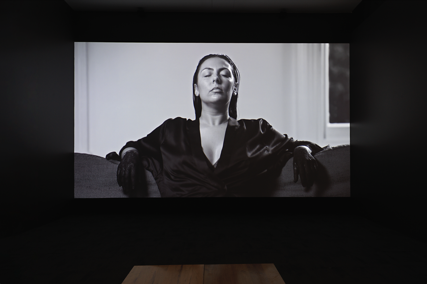 Monochromatic portrait of woman with eyes closed, wearing a robe and gloves, seated in front of a window and white wall.