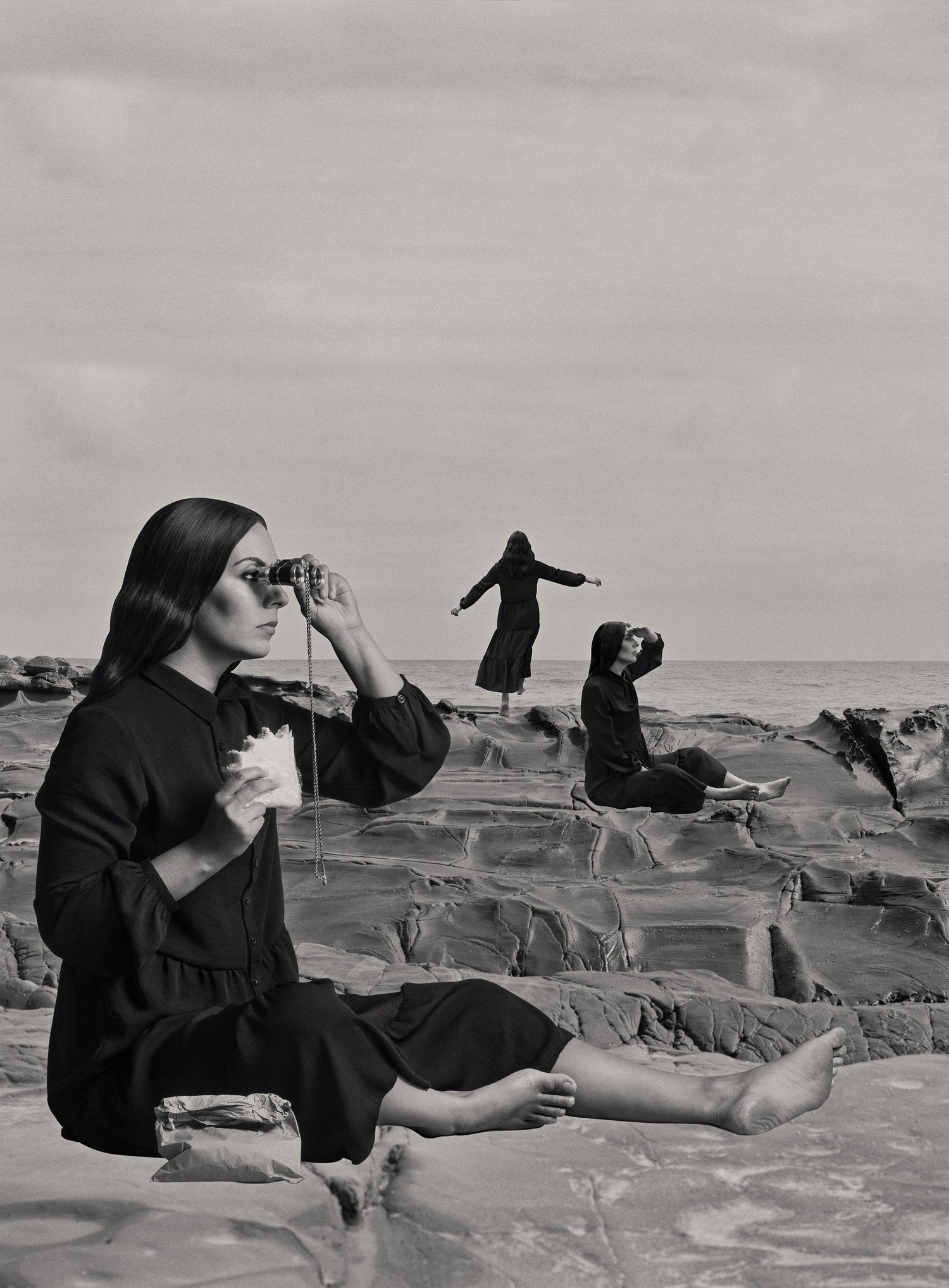 Three women on a rocky coast in a black and white photograph. One woman sits with binoculars, two other women in black clothing pose.