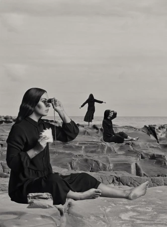Three women on a rocky coast in a black and white photograph. One woman sits with binoculars, two other women in black clothing pose.