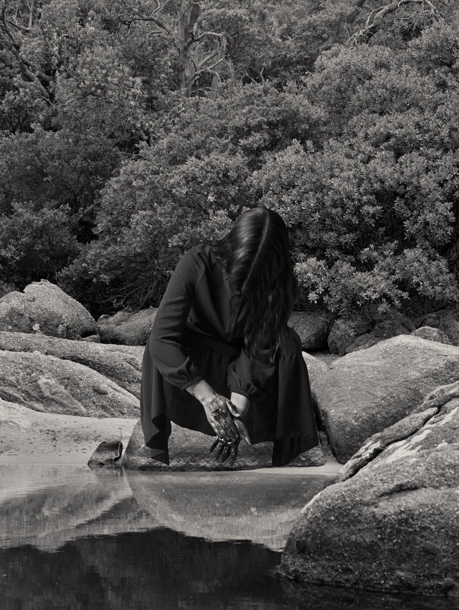 Monochrome photograph of a woman crouching by water amidst a rocky, wooded landscape with dense foliage and tree reflections.