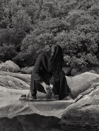 Monochrome photograph of a woman crouching by water amidst a rocky, wooded landscape with dense foliage and tree reflections.