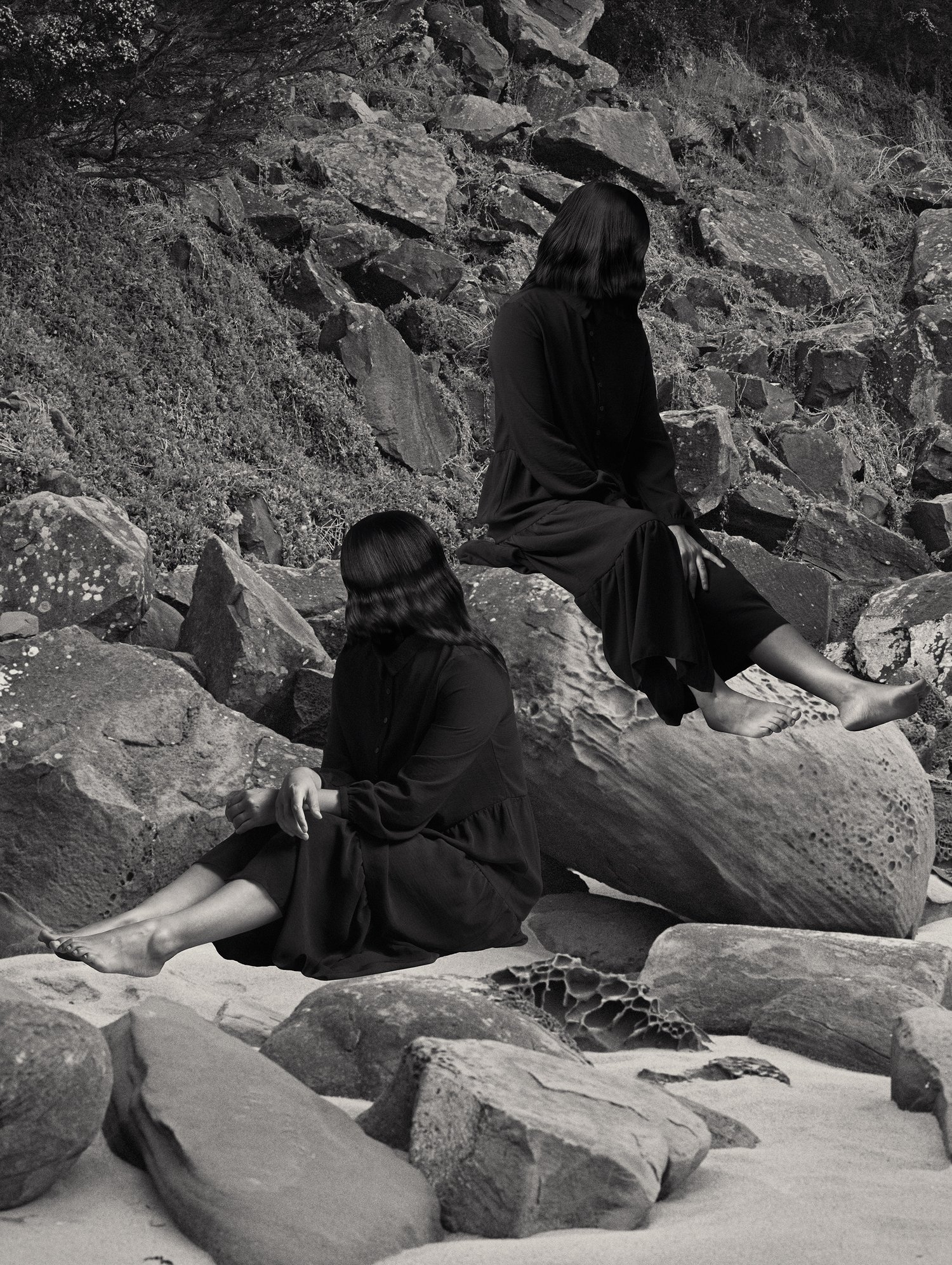 Two women in long black dresses sit among rocks on a beach, faces obscured by hair. Black and white, subdued lighting, overcast sky.