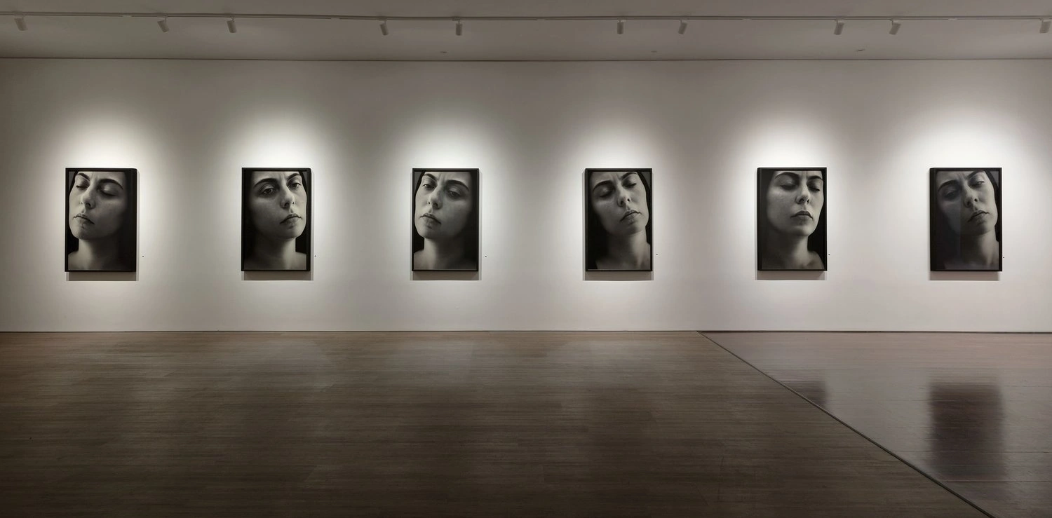 Six framed black and white portrait photographs hung in a row along a white gallery wall above a polished wooden floor.
