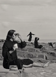 Three women in black dresses on rocky coast, one in foreground holding binoculars and a sandwich, by Hayley Millar Baker