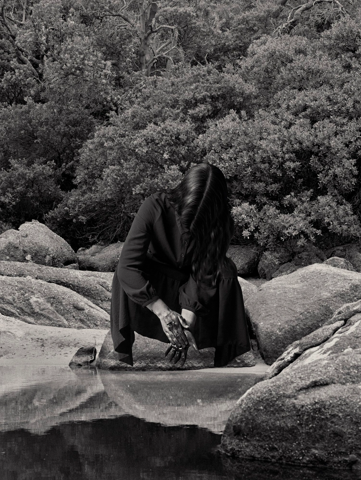 Woman in dark dress crouching by rocky water edge with dense foliage background in monochrome photography