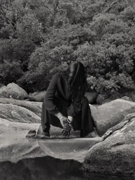 Woman in dark dress crouching by rocky water edge with dense foliage background in monochrome photography