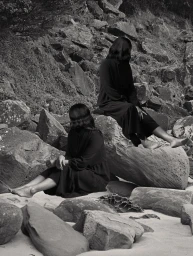 Monochrome photograph of two women with dark hair sitting on large boulders on a sandy beach by photographer Hayley Millar Baker