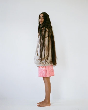 Profile view of a woman with long dark wavy hair wearing a beige zip-up hoodie and pink shorts against a plain white wall