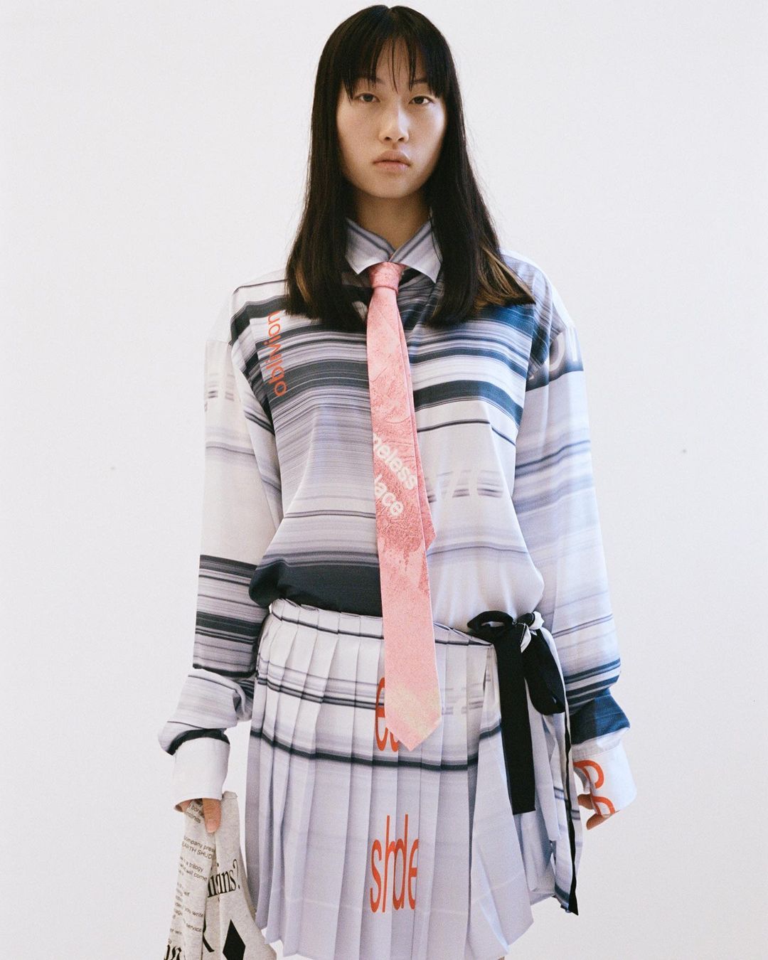 Young Asian woman in a white and blue striped shirt and pleated skirt with a pink tie, standing indoors against a white backdrop.