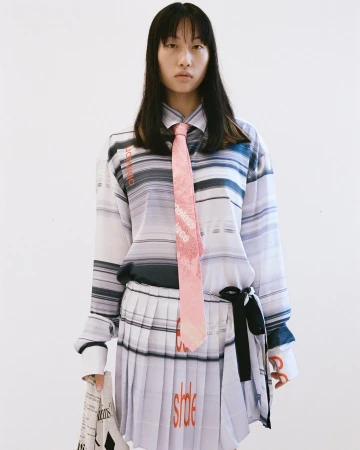 Young Asian woman in a white and blue striped shirt and pleated skirt with a pink tie, standing indoors against a white backdrop.