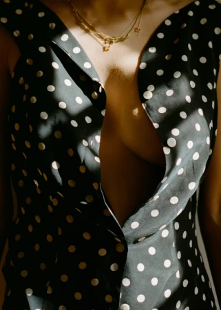 A woman's torso is partially visible, wearing a black polka-dot dress and gold necklaces, with shadows and light adding contrast.