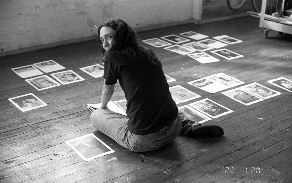 Woman sits on a wooden floor surrounded by scattered prints, looking back with a neutral expression in a black and white indoor scene.