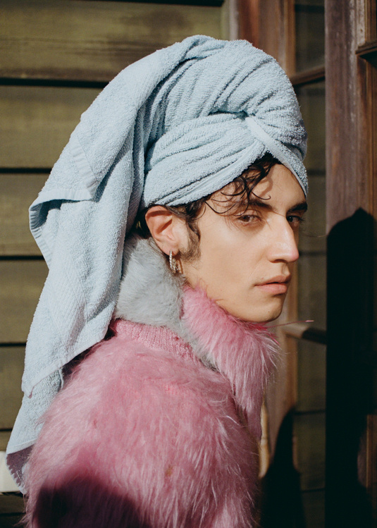 Portrait of a person with a towel wrapped around their head, posing in front of a wooden wall, wearing a fuzzy pink sweater.