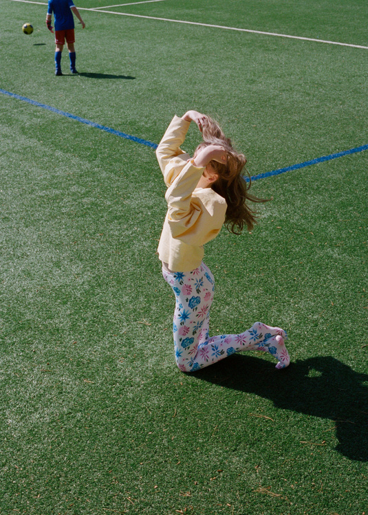 Young girl on knees with arms raised outdoors on green field, soccer player in background with ball on sunny day