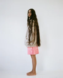 Profile view of a woman with long dark wavy hair wearing a beige zip-up hoodie and pink shorts against a plain white wall