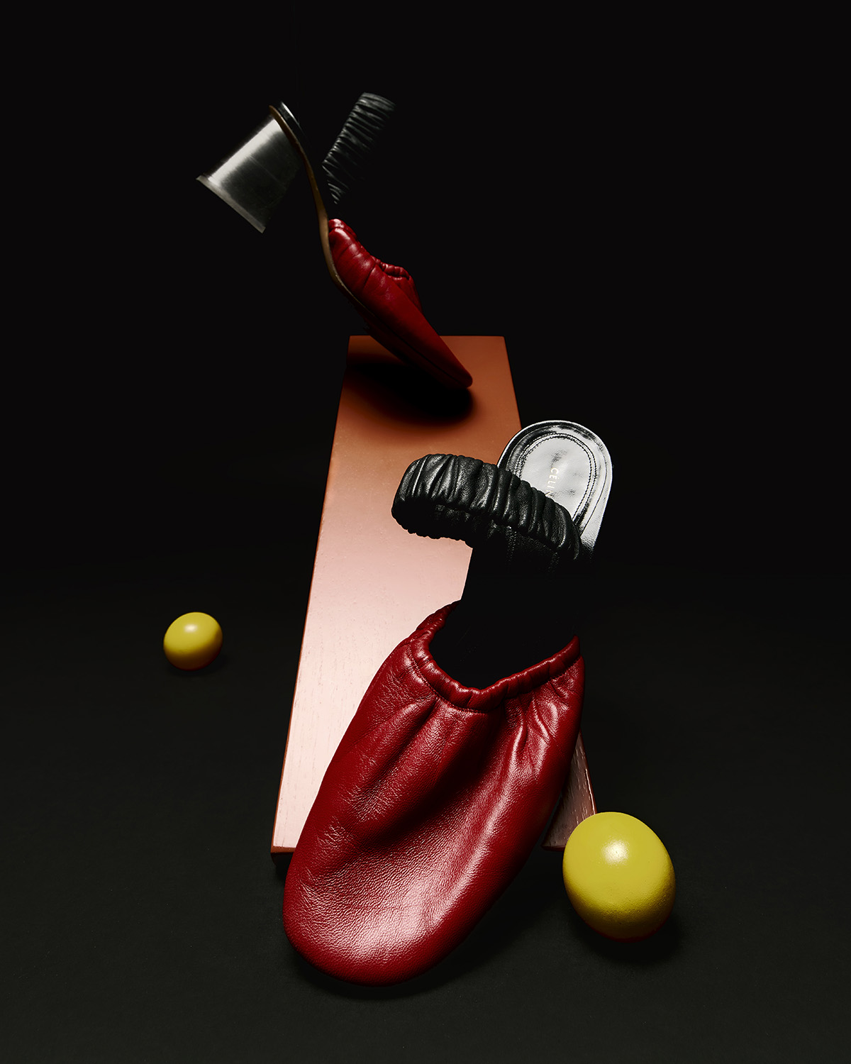 Fashion still life featuring a pair of red leather mule shoes, one balanced on a copper-colored block, against a dark background.