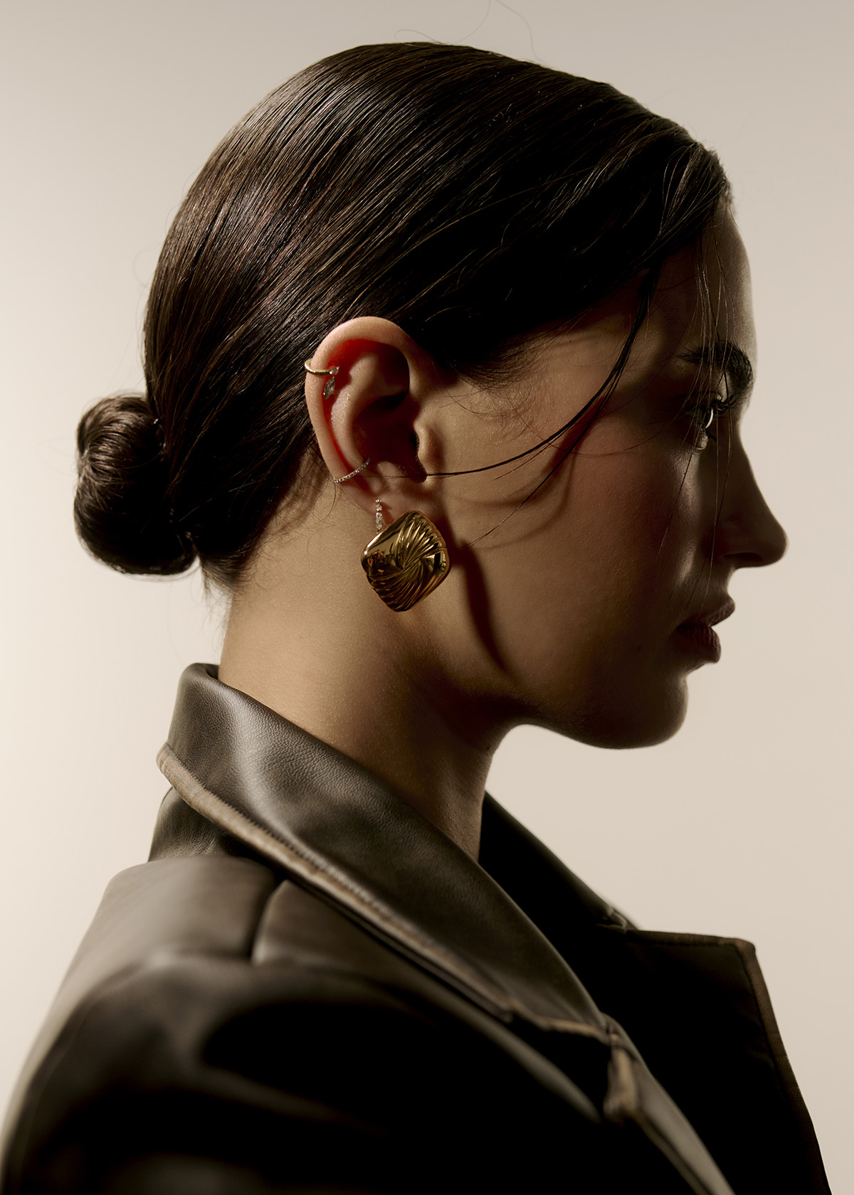 Profile portrait of a young woman with gold earrings and ear cuff, wearing a black leather jacket against a neutral background.