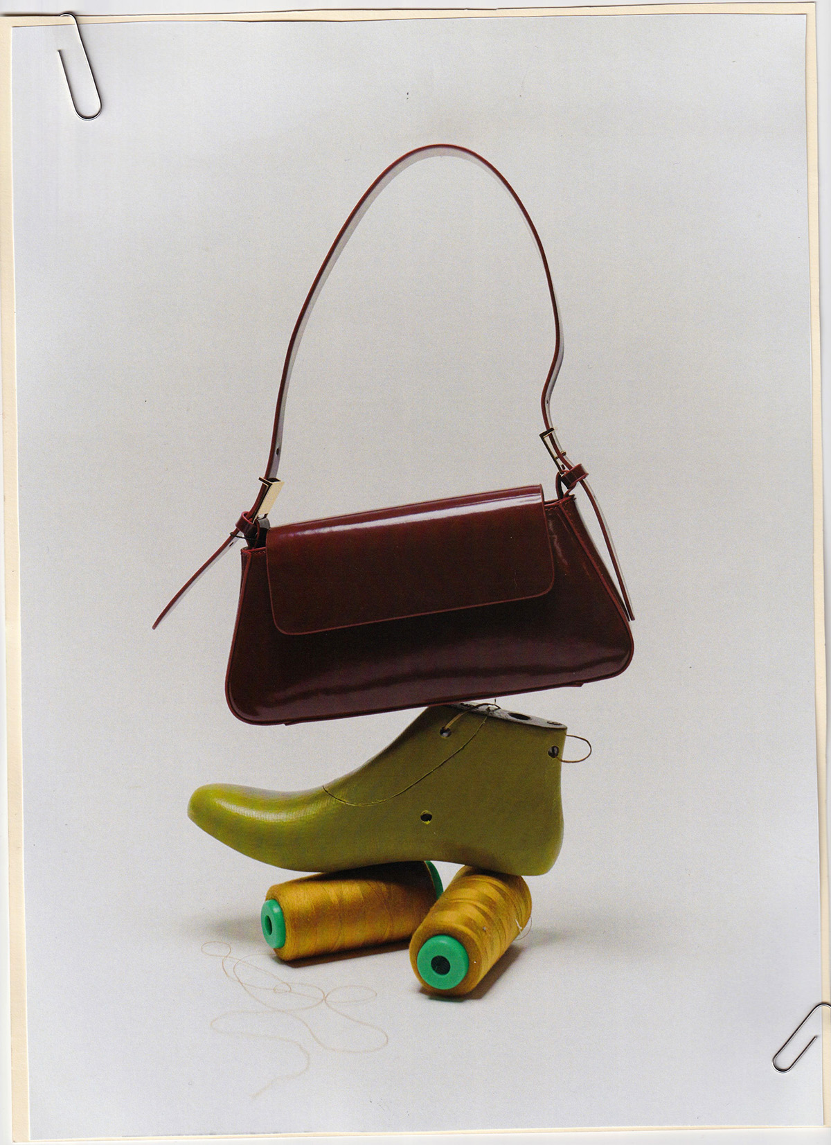 Still life of a burgundy purse atop a green shoe mold and two spools of gold thread, secured to a white board with metal clips.